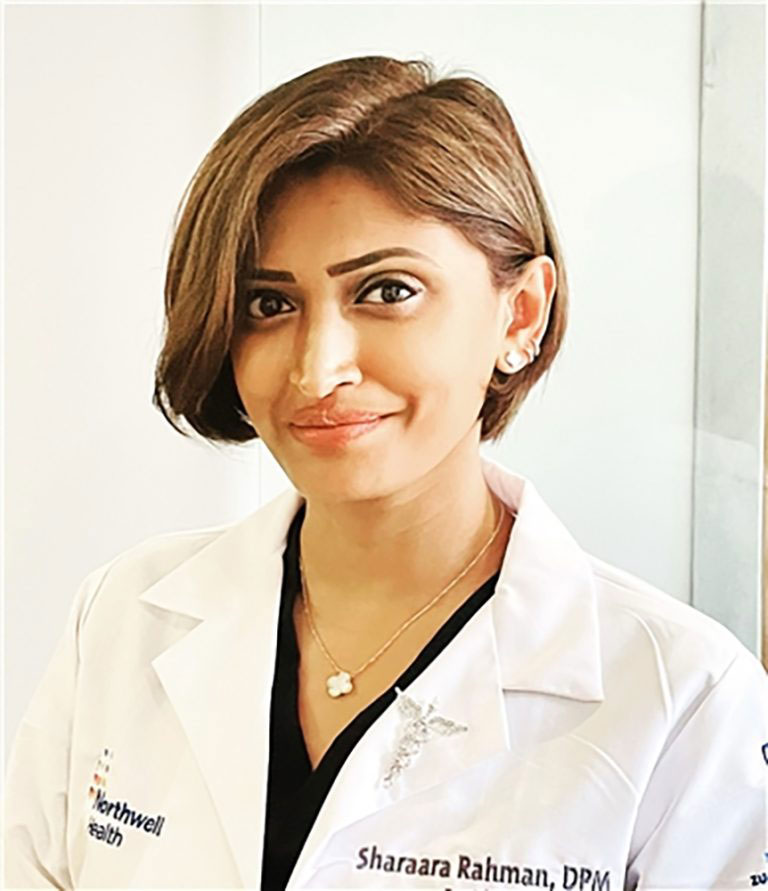 The image shows a person posing for the photograph. They are wearing a white lab coat, which suggests they might be a medical professional, and are smiling at the camera.