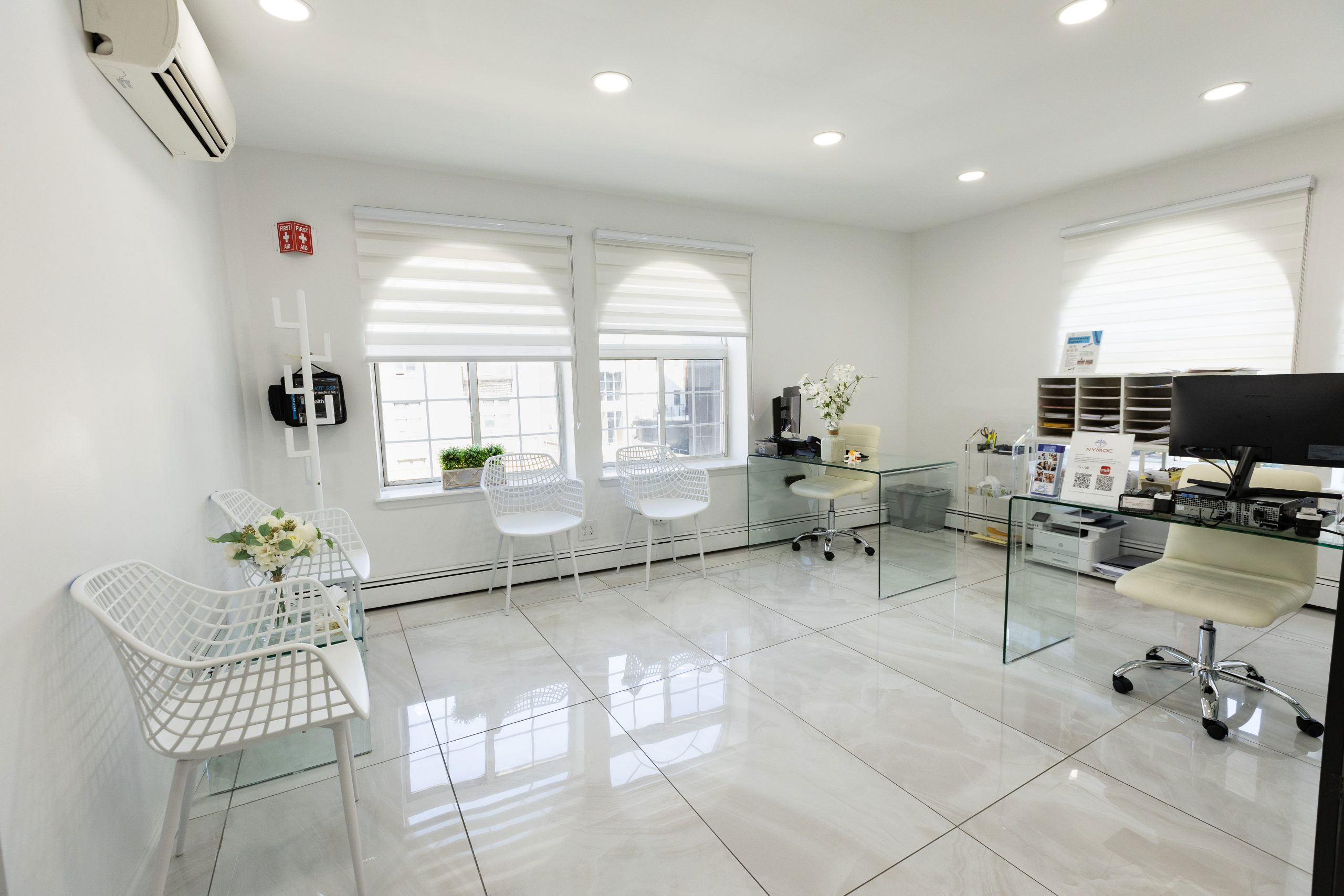 The image shows an interior space that appears to be a modern office or studio, featuring a clean and minimalist design with white walls, shiny flooring, a large window allowing natural light, a desk with a computer monitor, a chair, a glass table, a white couch, and various other decorative items.