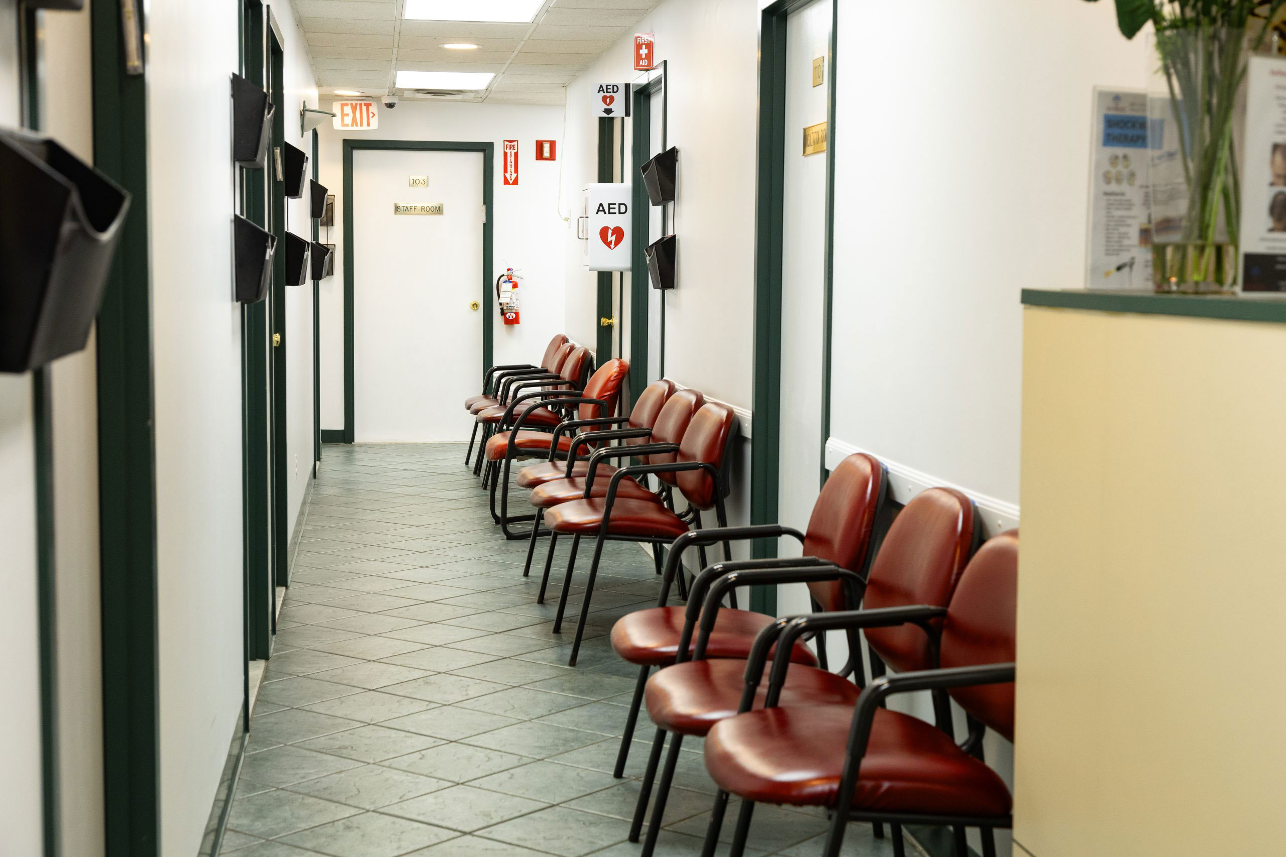 The image shows an interior hallway with a row of chairs, a long counter, and a door at the end.