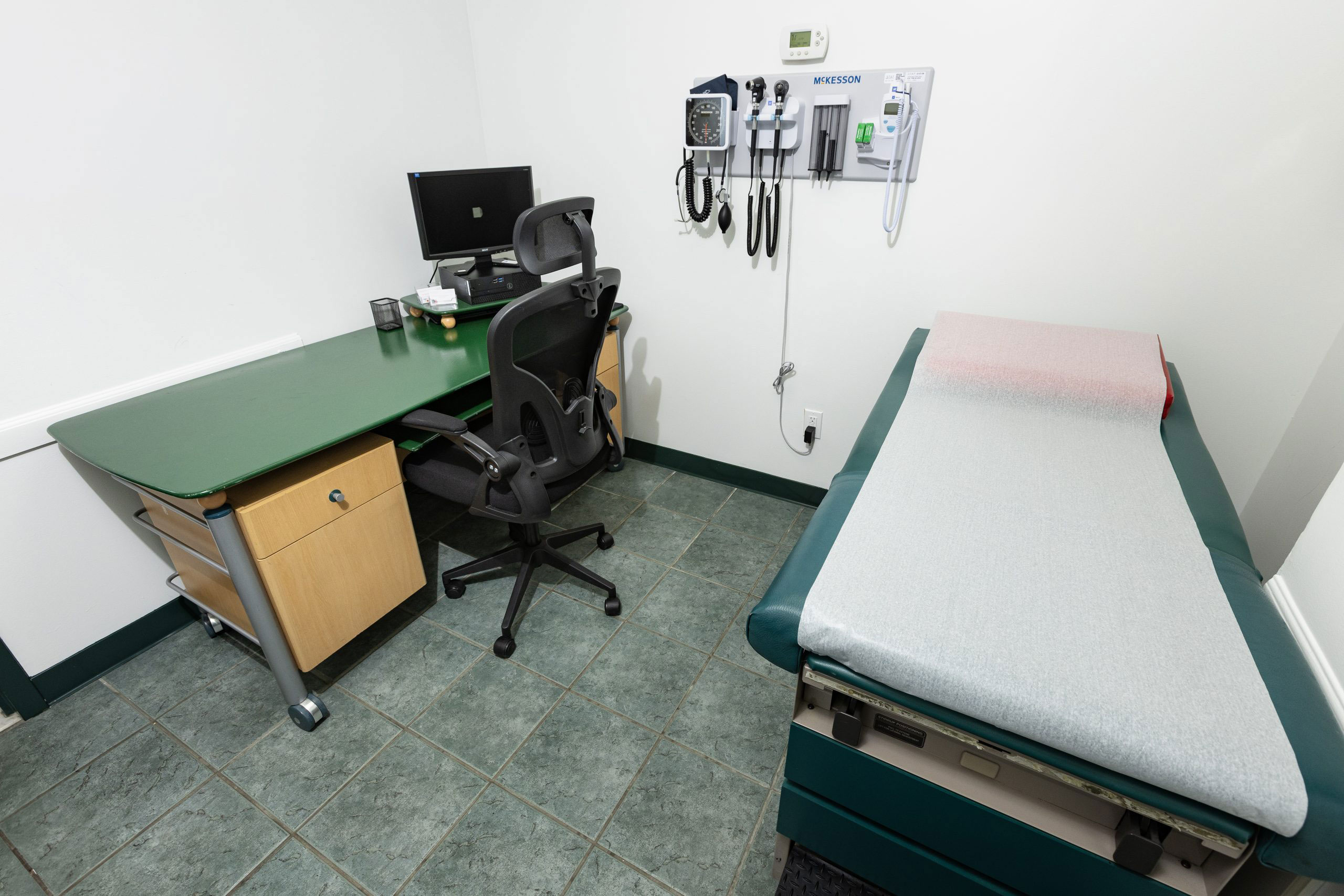 The image shows a small, well-equipped medical examination room with a desk and chair, a medical table, and various medical equipment.