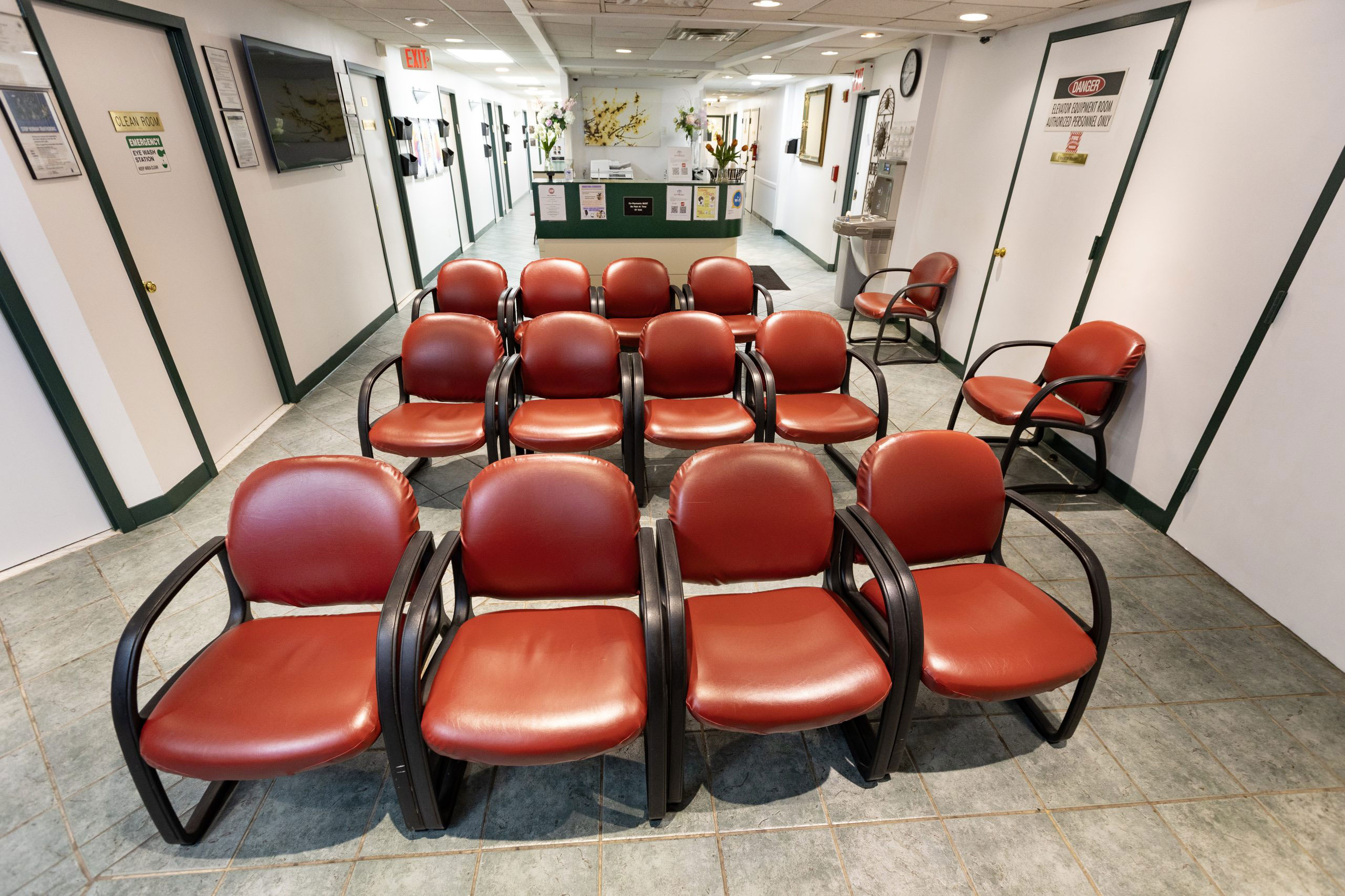 An image depicts an interior space with a row of red chairs on one side and a waiting area with a television mounted on the wall.