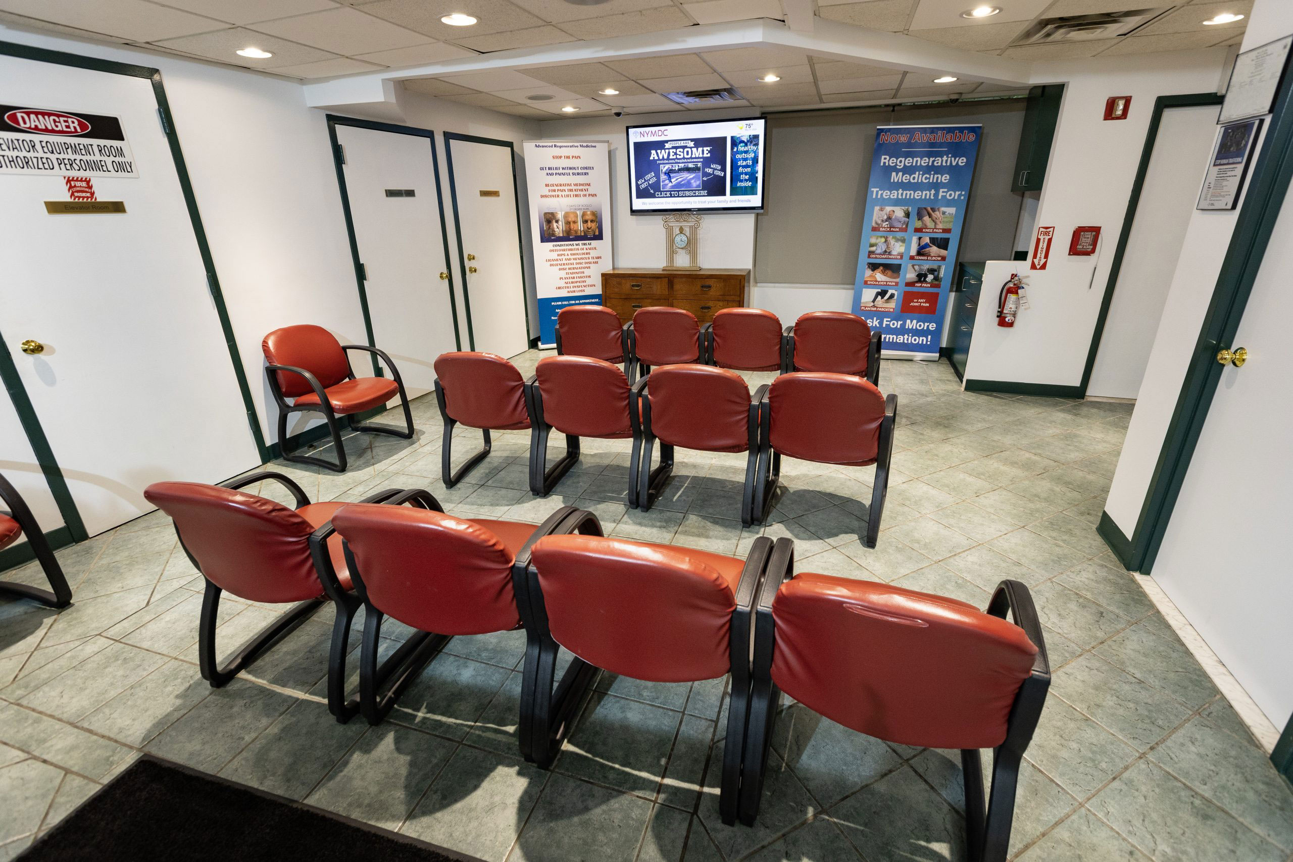 The image displays an interior space resembling a waiting room or small lobby, featuring chairs arranged in rows facing a central area with a television screen mounted on the wall. There is signage and a reception desk visible in the background.