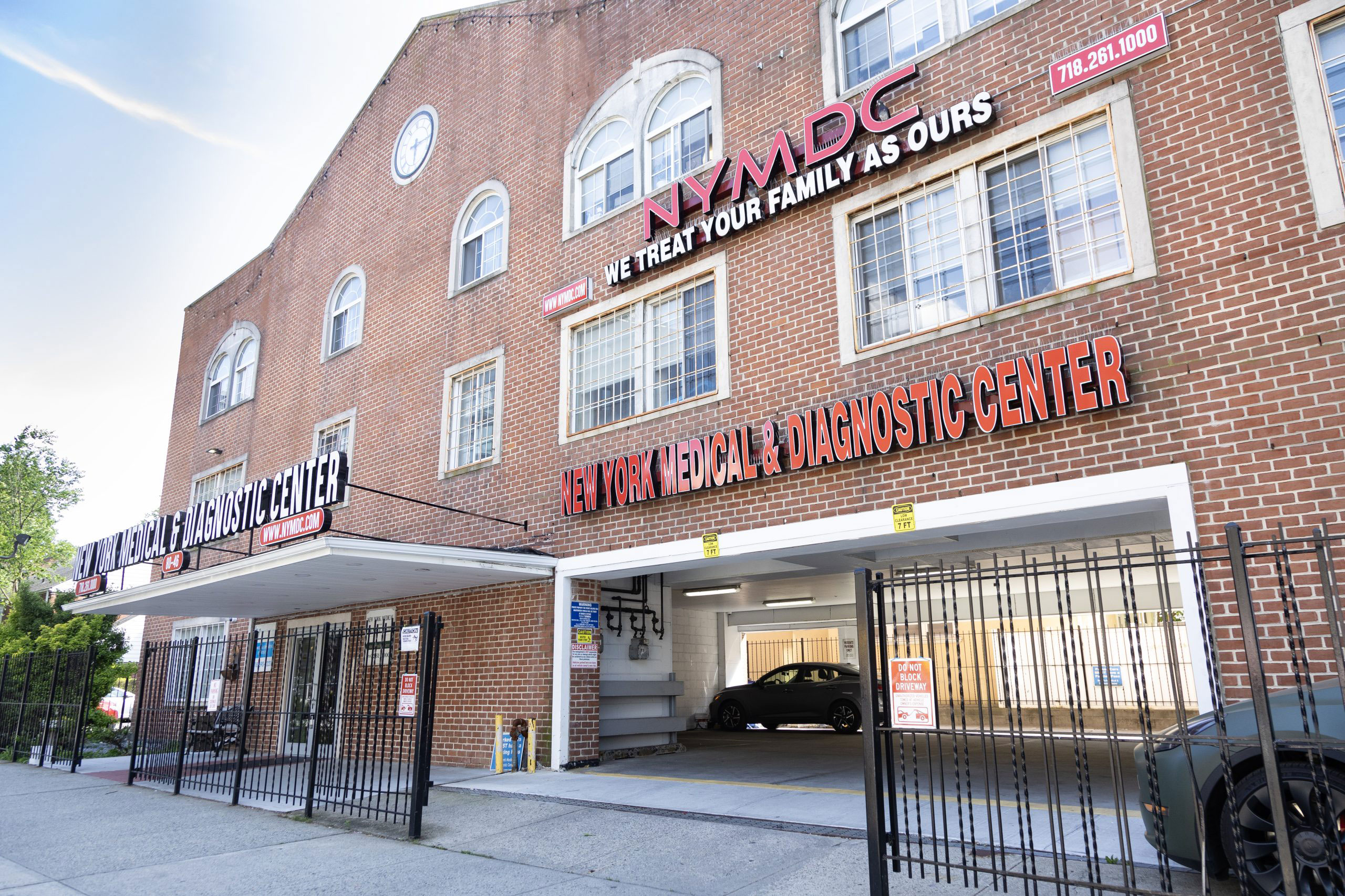 The image depicts a building with signage that includes the text 'NYMC,' 'WELCOME TO OUR FAMILY,' and other promotional information. It appears to be a medical center or healthcare facility, as indicated by the presence of a sign for the diagnostic center and 'NYMC' which could refer to New York Medical College. The building has a brick facade with a large window on the ground floor, and there is a parking lot in front with a gate that is currently open.