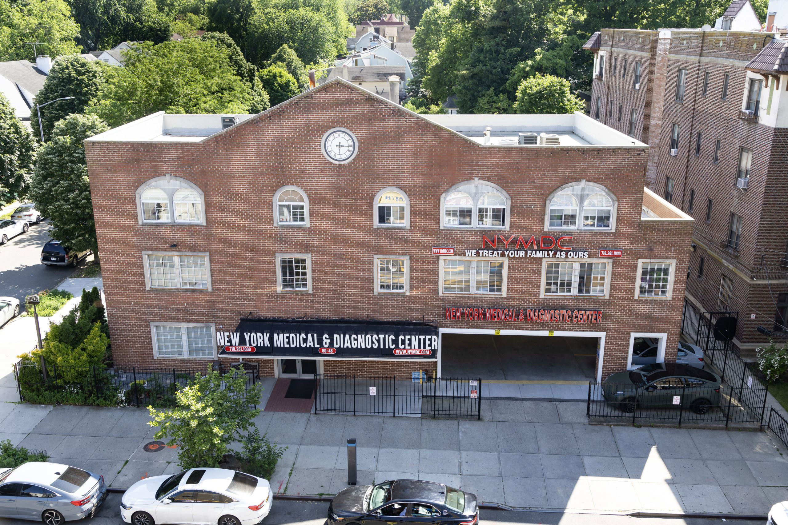 The image shows a two-story brick building with a sign that reads 'New York Medical Associates' and a clock on the front facade. It is situated in an urban area, as indicated by the presence of parked cars along the street and trees in the background.
