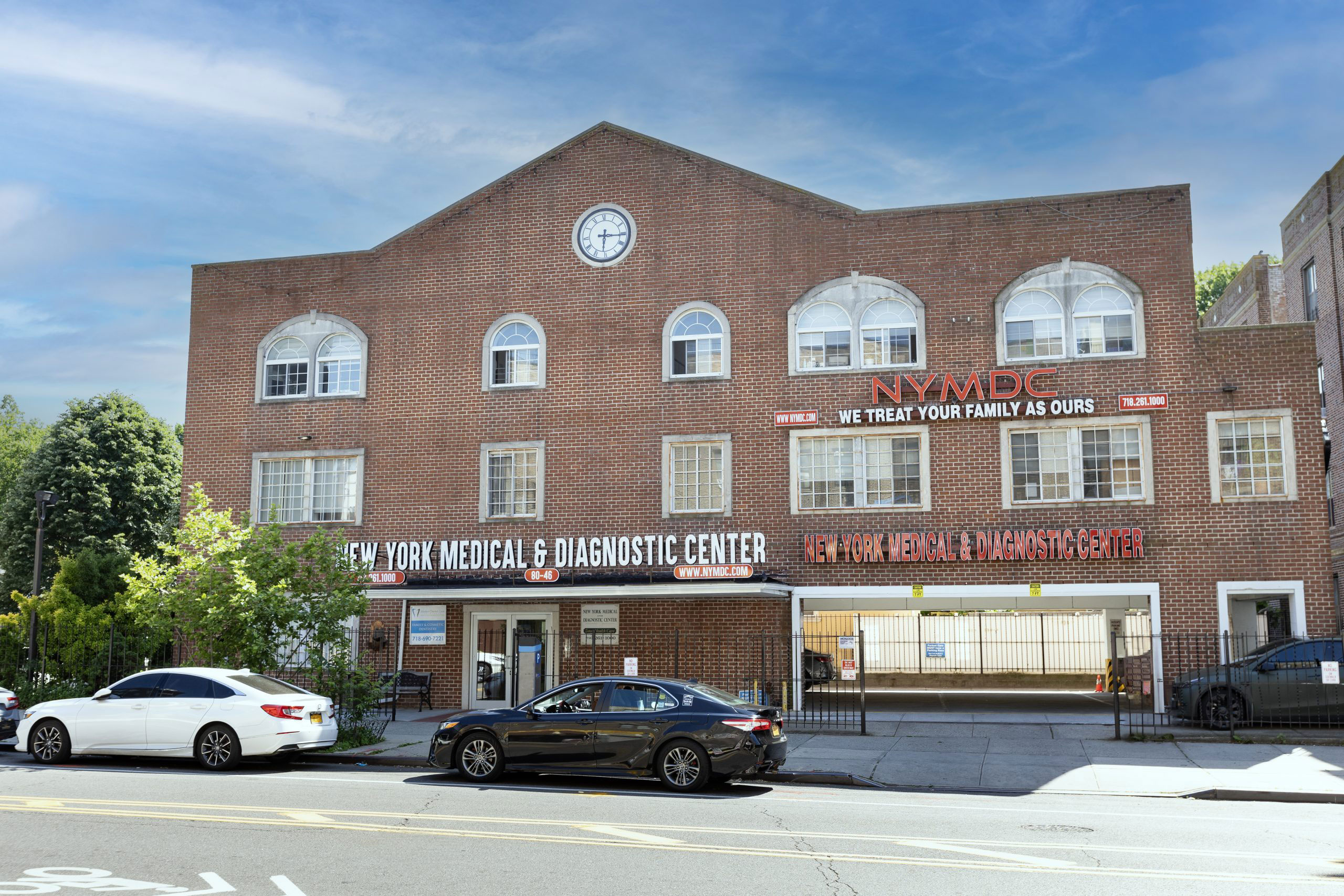 The image shows a two-story brick building with an arched entrance, featuring multiple signs and a clock on the facade. It is situated in an urban setting with trees and cars visible in the foreground.